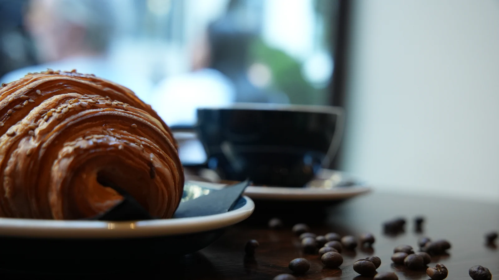 A golden, flaky croissant on a plate beside a dark cup of coffee and scattered beans